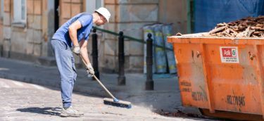 Warsaw, Poland - August 22, 2018: Old or new town street with man worker cleaner in blue uniform cleaning trash dust from cobblestone cobbled road by dumpster and sweeper tool during day